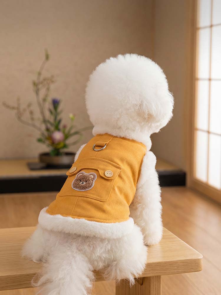 木の椅子に座る小型犬の後ろ姿。ビタミンイエローのデニム地に施されたクマの刺繍ポケットと、リードを繋ぐ丈夫なDカンが特徴。裾から覗く白いもこもこのボアが暖かさを演出し、和室の背景に溶け込む上品なデザイン。