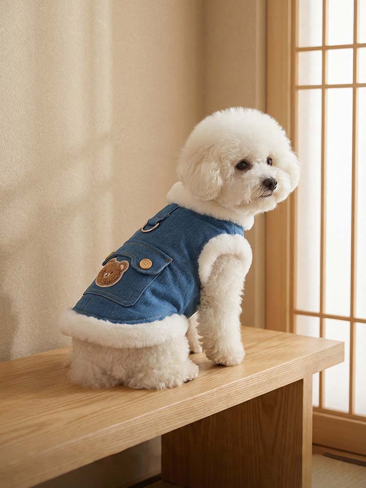 木製のベンチに座る、デニムブルーの裏ボアベストを着た小型犬。背中のDカンやボタン付きポケット、愛らしいクマの刺繍といったディテールが際立ち、首元の厚手ボアが冬の冷え込みから愛犬を優しく守るデザイン。