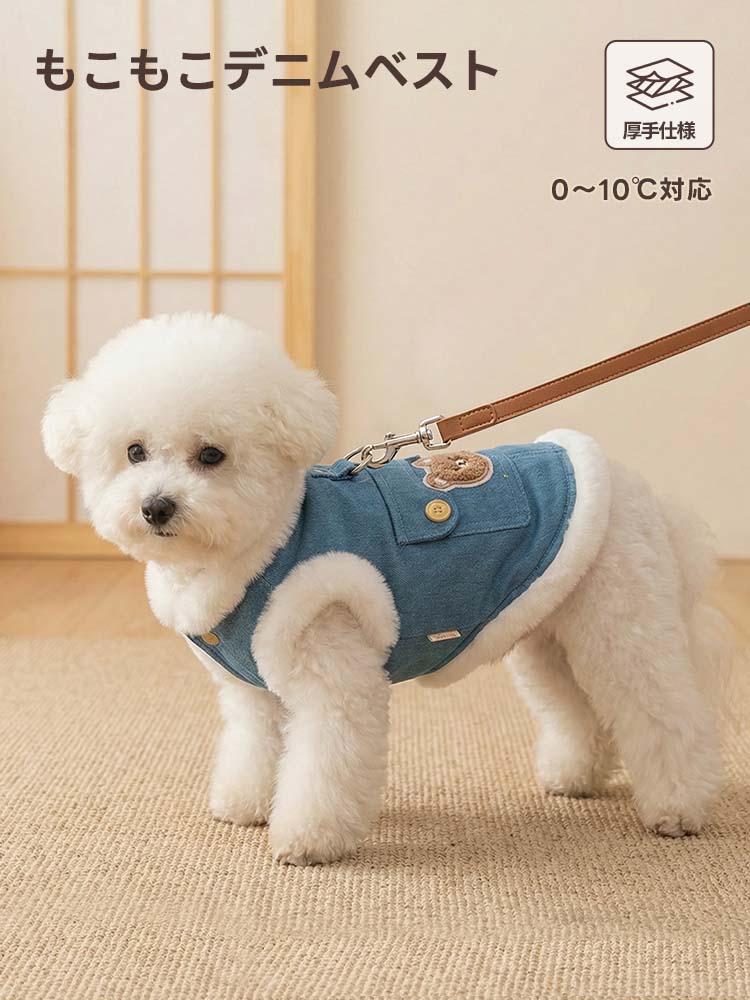 室内でリードを繋いだ白い犬が、厚手仕様のデニムベストを着て立つ姿。明るいブルーと白いボアのコントラストが際立ち、横から見てもシルエットが美しい冬用ペット服。背中のクマ刺繍とボタン付きポケットがアクセント。