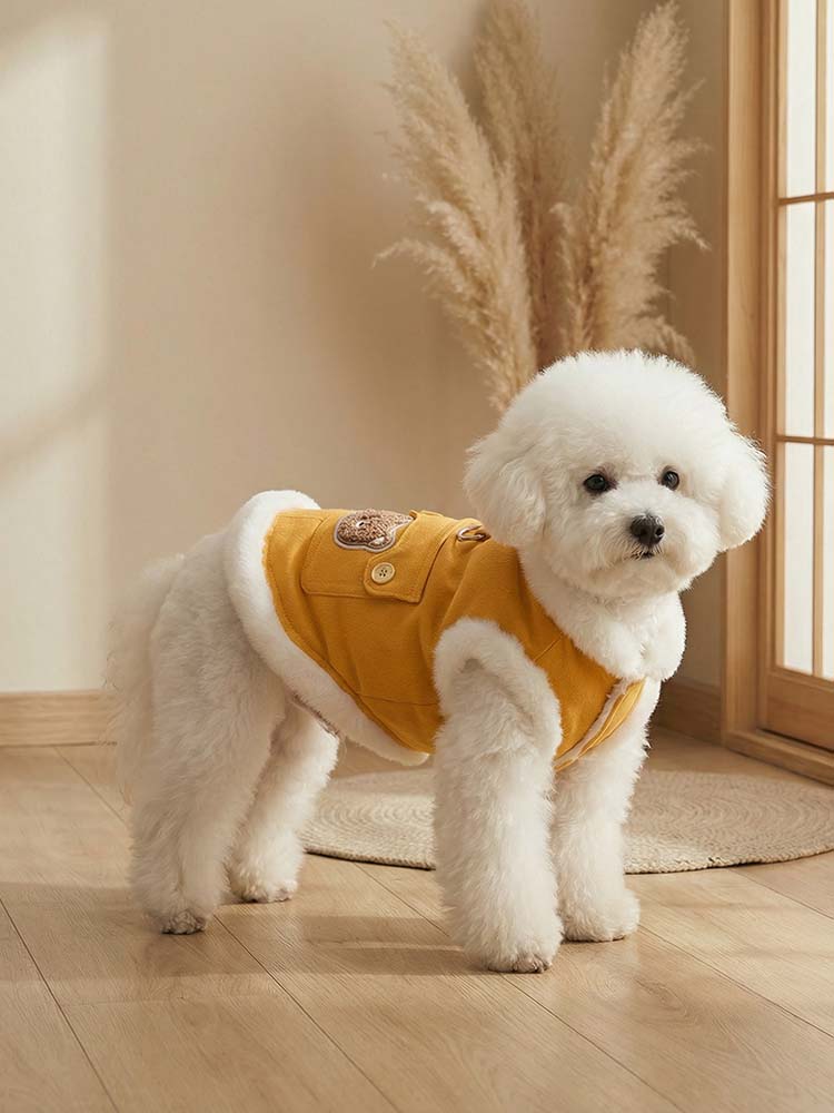 柔らかな光が差し込む室内で、ビタミンイエローの厚手デニムベストを着て立つ白い小型犬。首元や腕回りを包む白い裏ボアと黄色のコントラストが美しく、背中のクマ刺繍とリードフックが映えるお洒落な冬用ペットウェア。